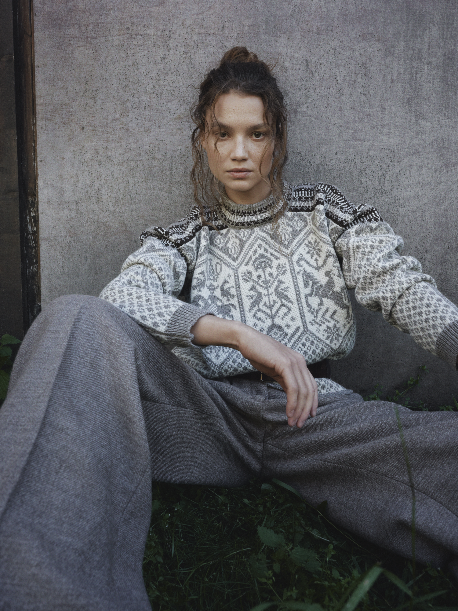 Modell iført ufarget 1994 Lillehammer ullgenser for dame fra Dale of Norway, i tradisjonelt mønster i grå beige toner, stylet med vide ullbukser foran en betongvegg