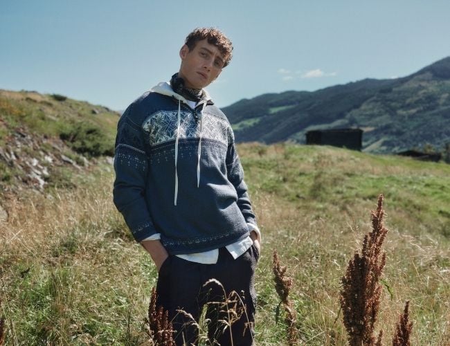 Ung mann i ullgenser med tradisjonelt mønster fra Dale of Norway, stående i grønt fjellandskap med blå himmel i bakgrunnen