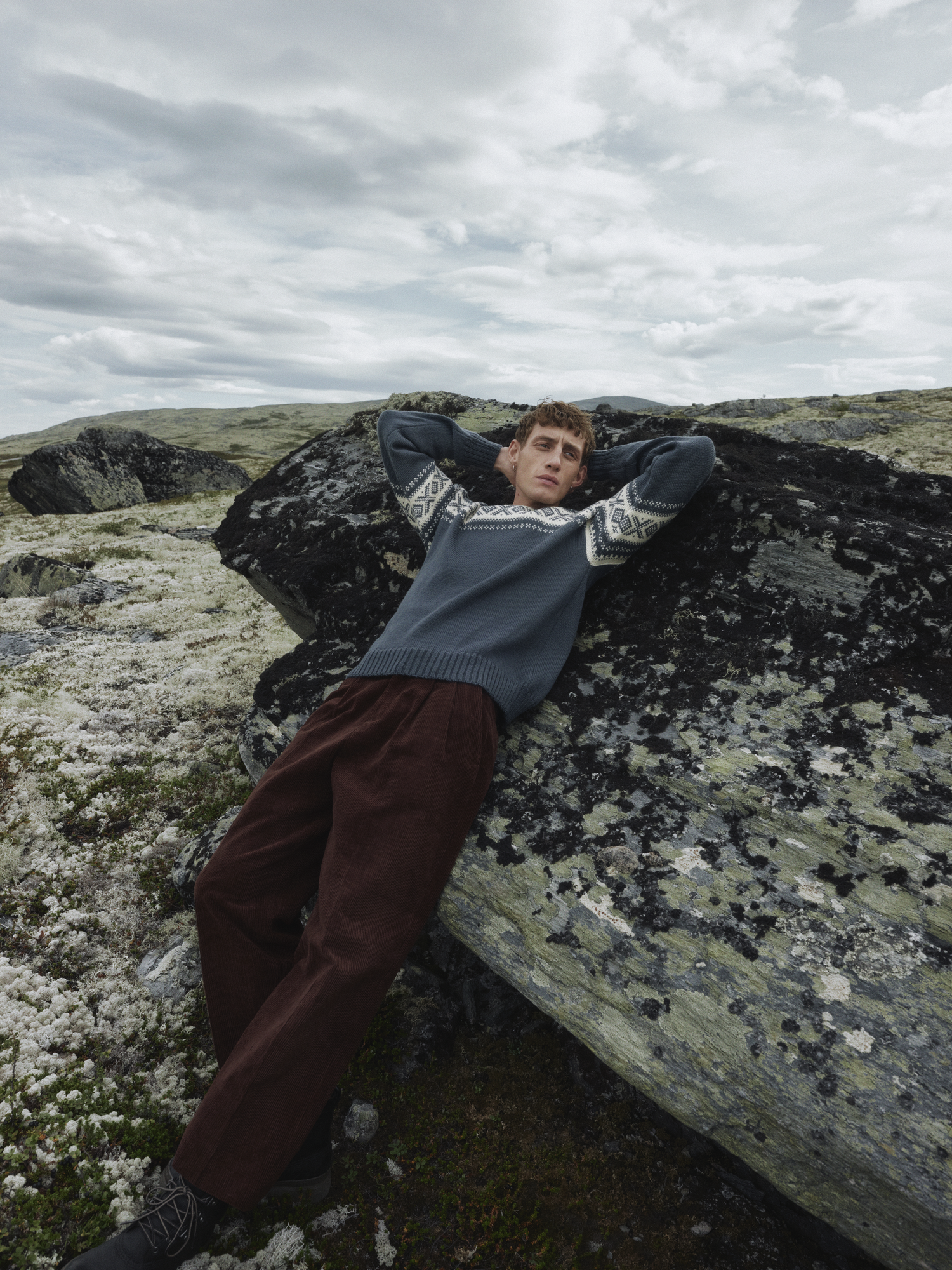 Modell i Cortina 1956 Unisex genser fra Dale of Norway i stålblå og offwhite, liggende avslappet mot lavakledde fjell i høyfjellet under overskyet himmel