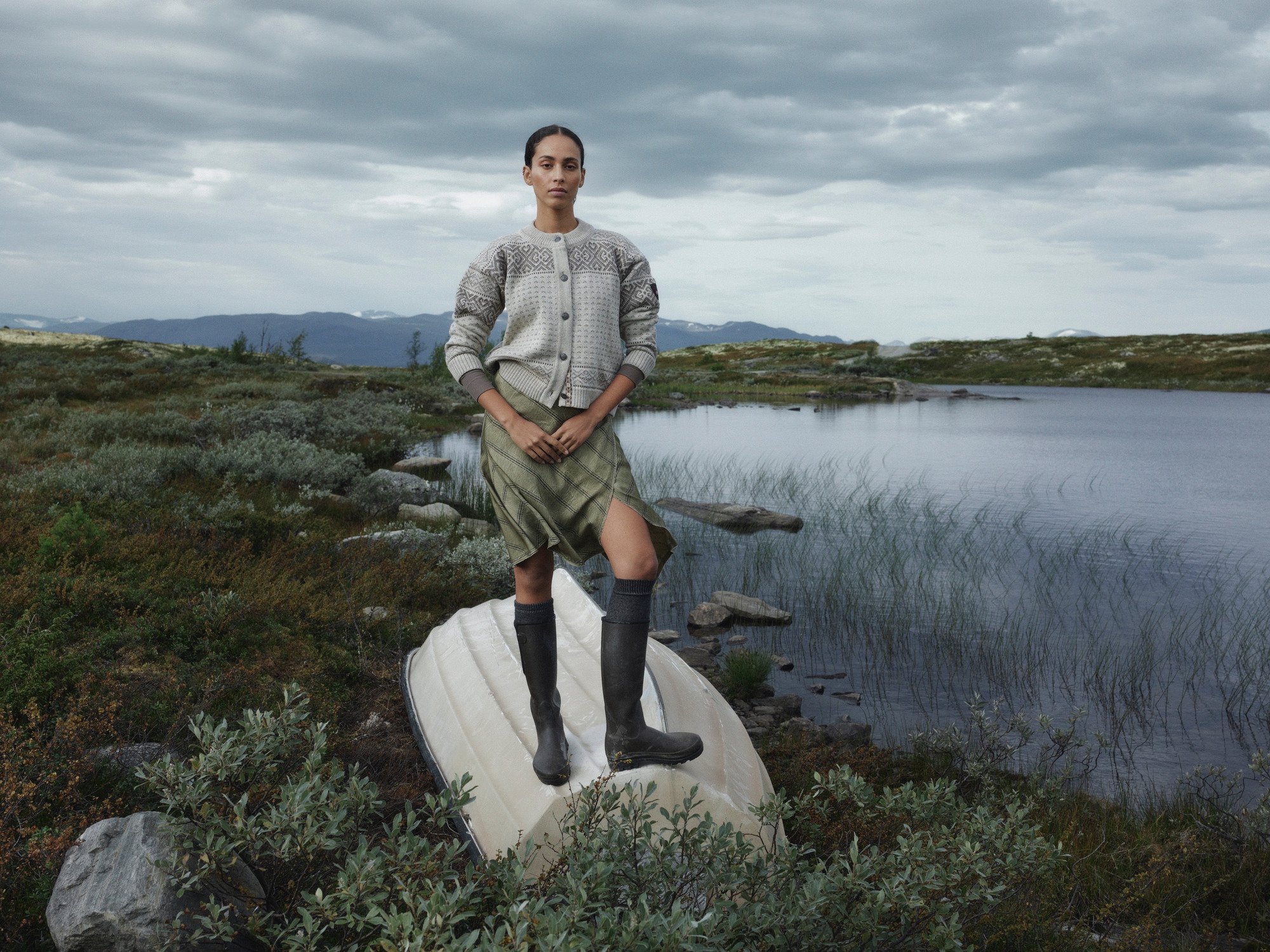 Modell iført Hannah cardigan fra Dale of Norway i naturfarget ull, stående på en stein ved fjellvann med dramatiske skyer og norsk høyfjellsnatur i bakgrunnen