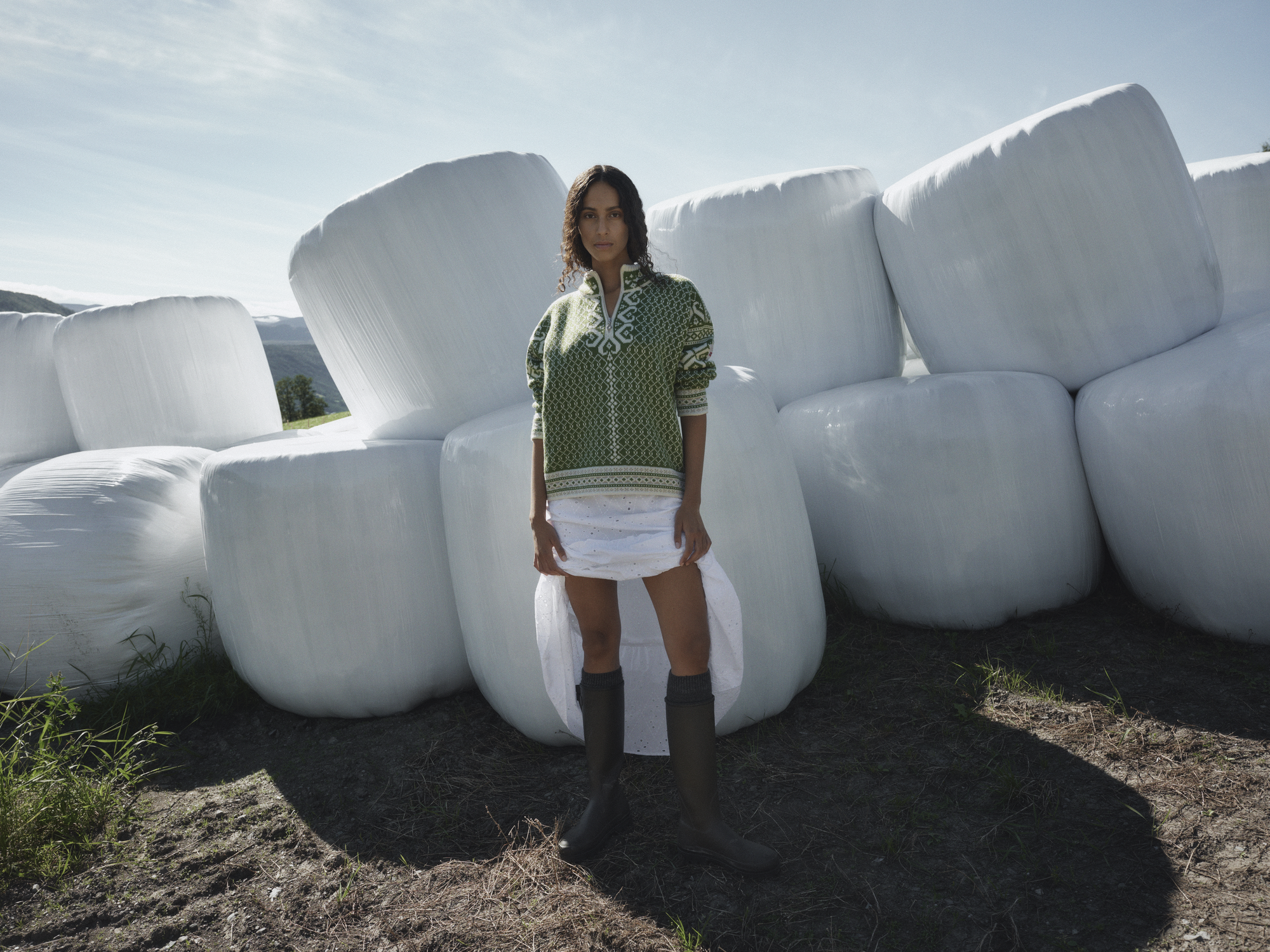 Leknes ullgenser for dame fra Dale of Norway i vårgrønt mønster, stylet med hvitt skjørt og boots foran store rundballer i norsk jordbrukslandskap