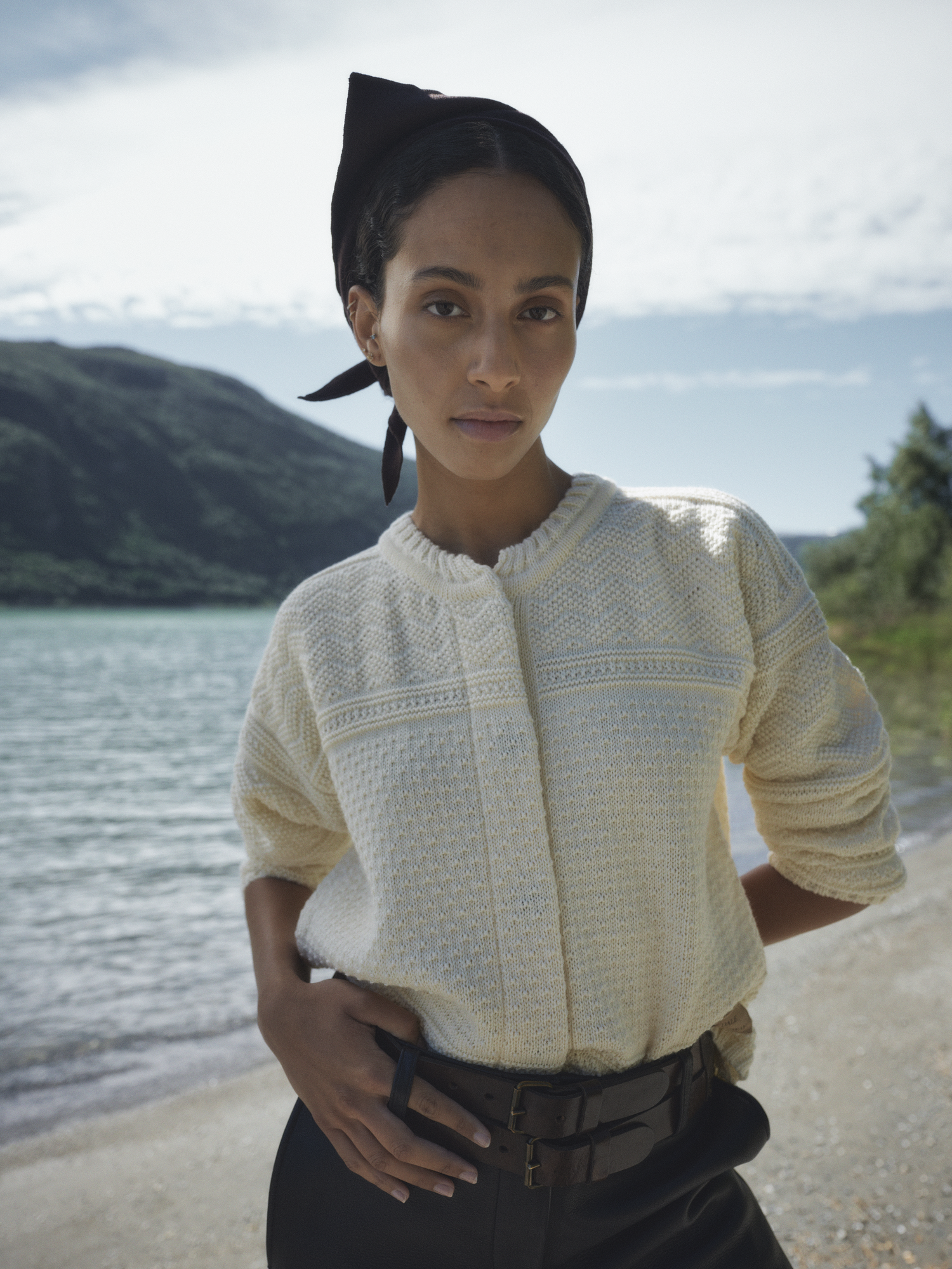 Modell iført Lusa cardigan for dame fra Dale of Norway i lys norsk ull, med belte i livet, fotografert ved vannkanten med fjell og blå himmel i bakgrunnen