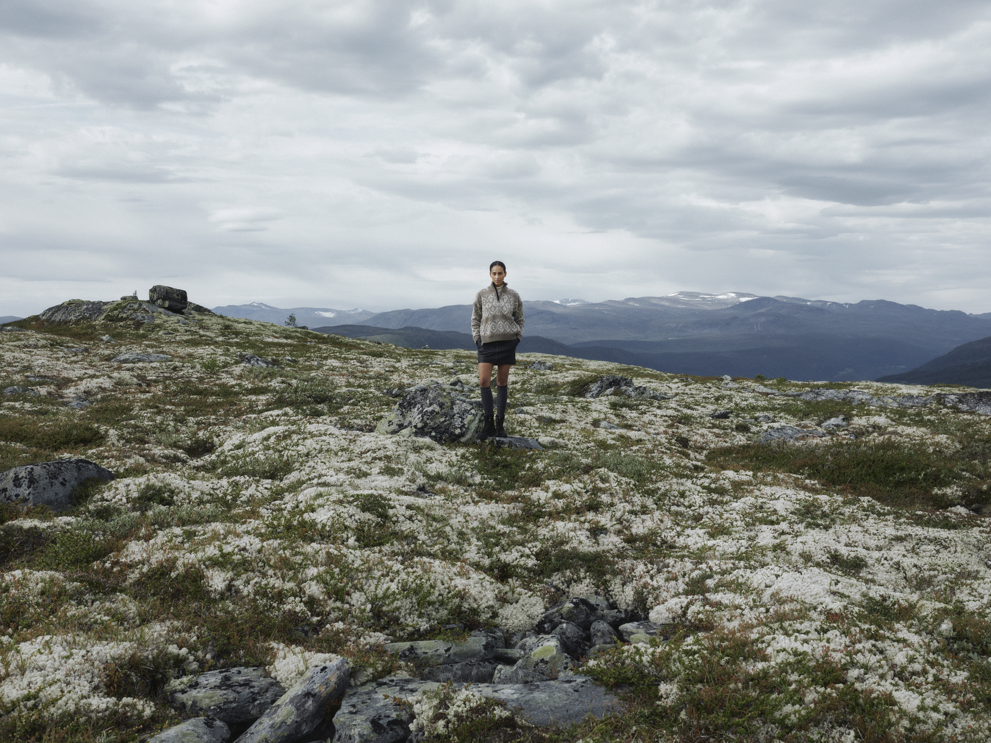 Modell i Roset vindtett ullgenser for dame fra Dale of Norway, stående alene i åpent høyfjellsterreng under dramatisk, overskyet himmel