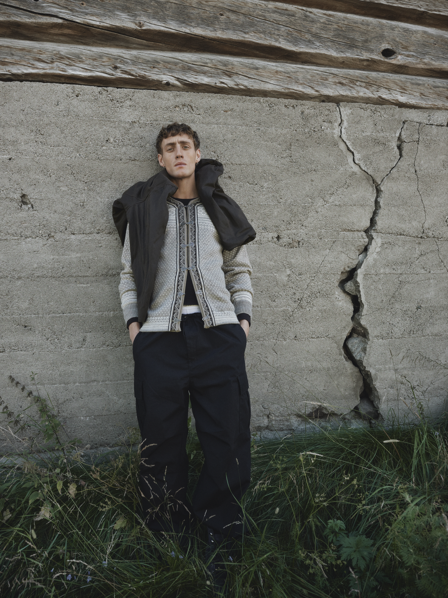 Modell i Setesdal unisex genser fra Dale of Norway i undyed ull, stylet med mørk bukse og jakke over skuldrene, foran en røff betongvegg med sprekker og gress