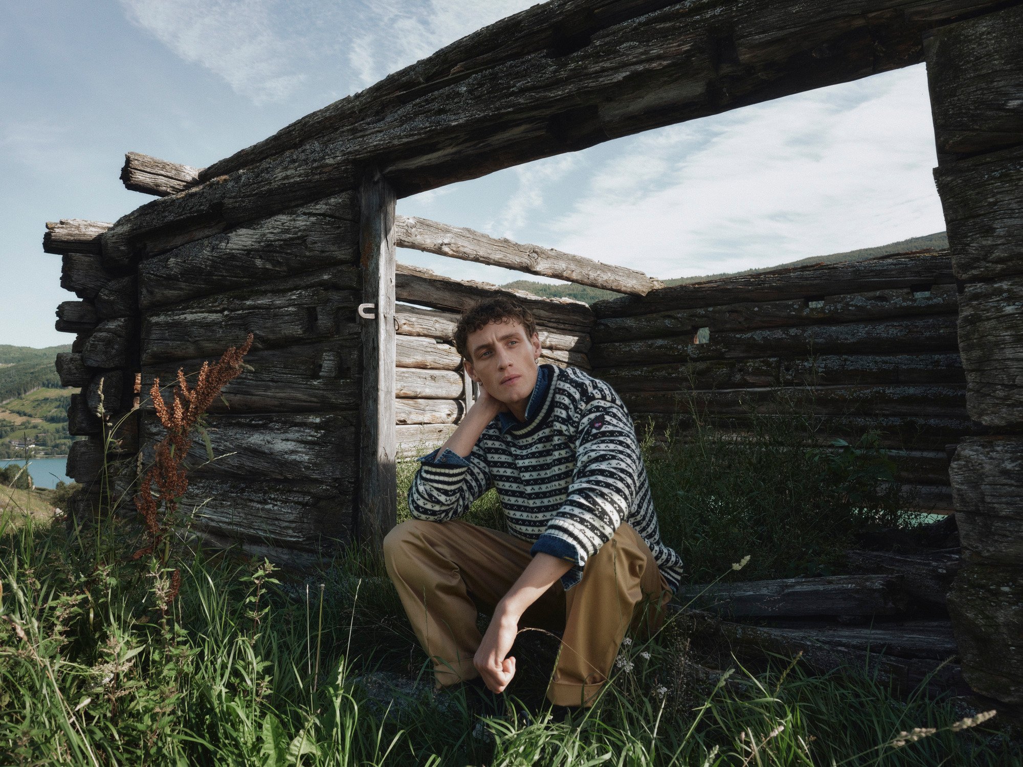 Skipsleia unisex ullgenser fra Dale of Norway, i norsk ull, brukt av modell foran en værbitt tømmerhytte i fjellandskap