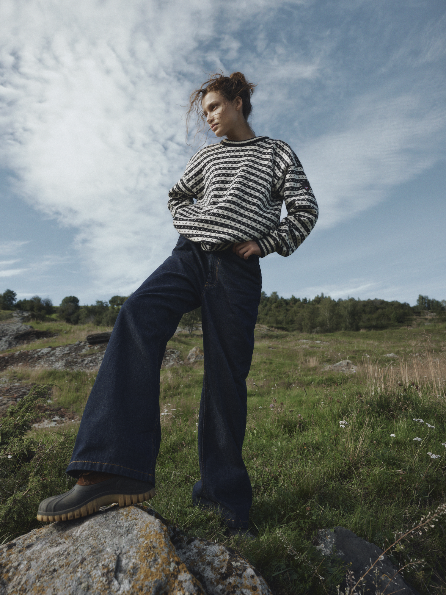 Modell iført Skipsleia unisex genser i norsk ull fra Dale of Norway, såtende på en stein i åpent grønt landskap med dramatisk himmel og vide jeans